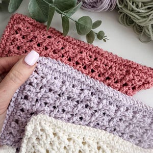 Puede incluir: Primer plano de tres pa&ntilde;os de ganchillo en tonos rosa, lavanda y crema. Los pa&ntilde;os tienen un tejido abierto y texturizado. Una mano con una u&ntilde;a pintada de rosa sostiene el pa&ntilde;o rosa. Madejas de hilo en colores coordinados en el fondo.