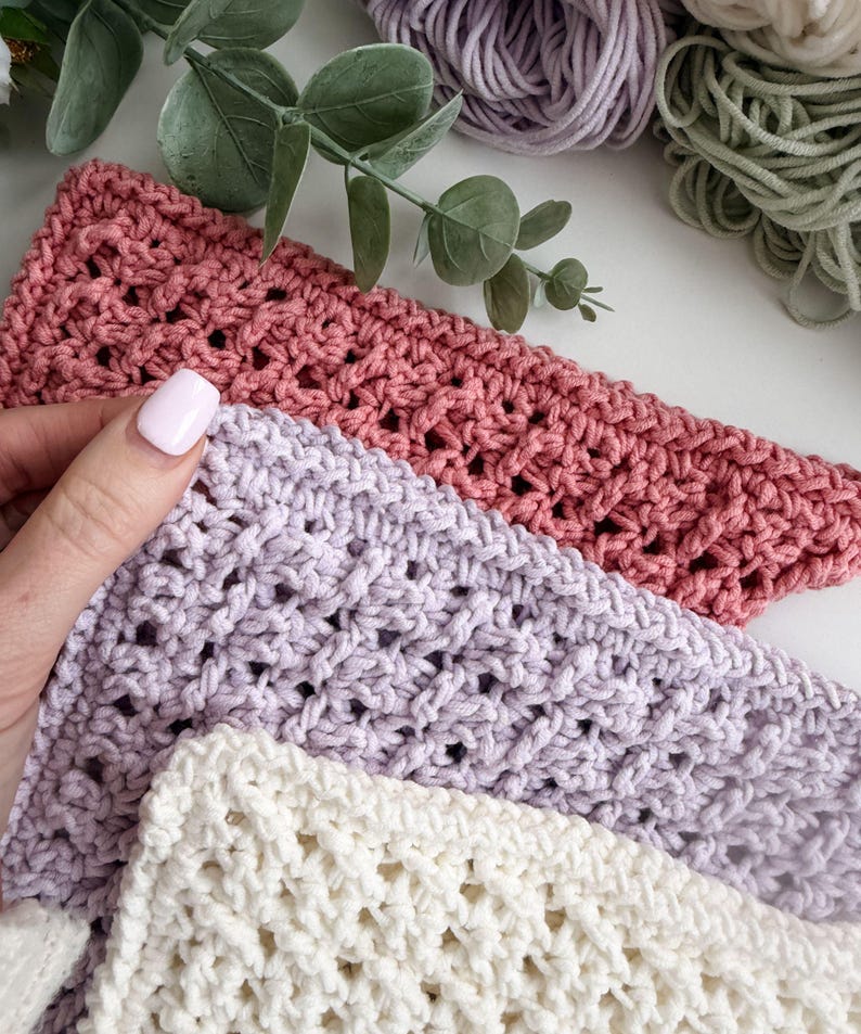 Puede incluir: Primer plano de tres pa&ntilde;os de ganchillo en tonos rosa, lavanda y crema. Los pa&ntilde;os tienen un tejido abierto y texturizado. Una mano con una u&ntilde;a pintada de rosa sostiene el pa&ntilde;o rosa. Madejas de hilo en colores coordinados en el fondo.