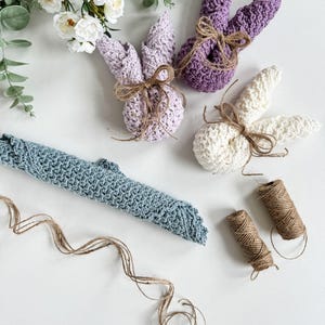May include: Three crocheted bunny shapes in purple, white, and blue, tied with twine. The bunnies are on a white surface with a roll of twine and a sprig of white flowers.