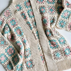 May include: A crocheted cardigan featuring a granny square pattern in shades of beige, pink, and teal. The cardigan has a long, open front and long sleeves.