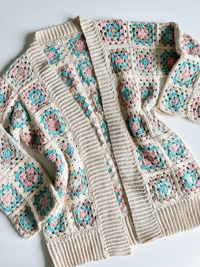 May include: A crocheted cardigan featuring a granny square pattern in shades of beige, pink, and teal. The cardigan has a long, open front and long sleeves.