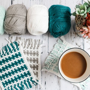 May include: Four balls of yarn in teal, white, gray, and mint green colors, with three crocheted coasters in teal, gray, and mint green colors, and a cup of coffee on a white wooden surface.
