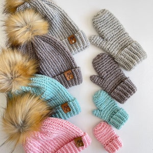 May include: Set of five crocheted hats and mittens in various colors, including gray, brown, blue, and pink. Each hat has a faux fur pom pom and a brown leather tag with the text "Mida's".