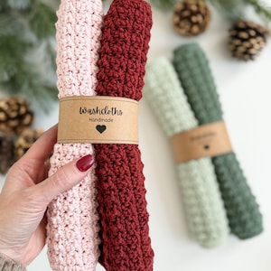 May include: Handmade washcloths in pink and burgundy, held together with a brown paper band. The band reads "Washcloths Handmade" with a heart symbol. Additional washcloths in green and light green are in the background.
