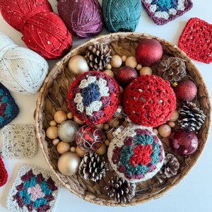 May include: A basket filled with crocheted baubles in red, white and green. The baubles are shaped like balls and have a variety of patterns. There are also some pine cones, wooden beads and red and white baubles in the basket.