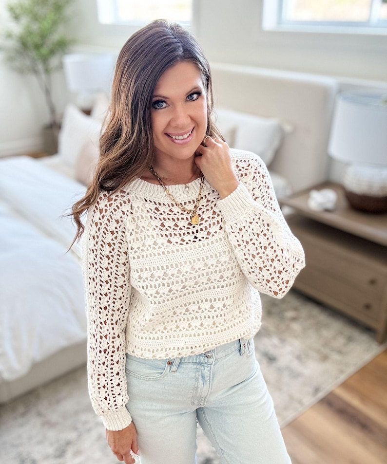 K&ouml;nnte beinhalten: Ein cremefarbener H&auml;kelpullover mit langen &Auml;rmeln und rundem Ausschnitt. Der Pullover hat ein offenes, luftiges Design. Die Person tr&auml;gt eine hellblaue Jeans und eine goldene Halskette.