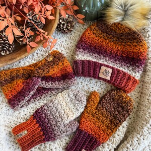 May include: A fall-themed crochet set featuring a beanie with a faux fur pom, a headband, and mittens. The set showcases a gradient of colors, including orange, burgundy, and cream. Each piece has a leather tag. The items are displayed on a cream-colored, textured surface.
