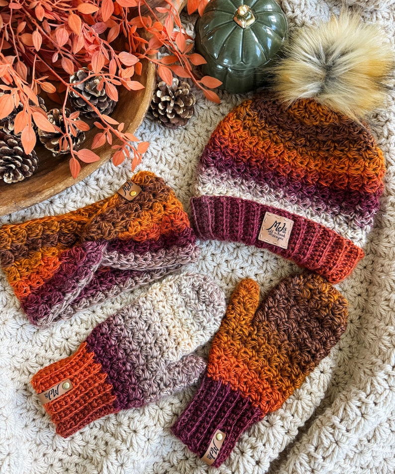 May include: A fall-themed crochet set featuring a beanie with a faux fur pom, a headband, and mittens. The set showcases a gradient of colors, including orange, burgundy, and cream. Each piece has a leather tag. The items are displayed on a cream-colored, textured surface.