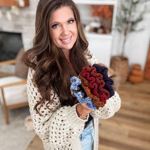 May include: A woman wearing a cream-colored, open-weave crochet cardigan holds a collection of colorful crochet items. The items include shades of blue, red, brown, and purple. The cardigan has a long, open design.