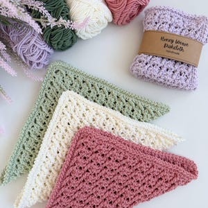 Puede incluir: Pa&ntilde;os de cocina de ganchillo hechos a mano en tonos rosa, crema y verde, junto con ovillos de lana a juego. Un pa&ntilde;o de cocina est&aacute; envuelto con una banda de papel marr&oacute;n que dice "Honey Weave Dishcloth Handmade".