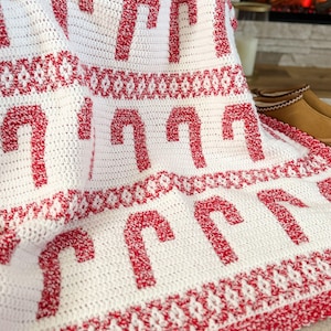 K&ouml;nnte beinhalten: Eine wei&szlig;e geh&auml;kelte Decke mit einem sich wiederholenden Muster aus roten Zuckerstangen. Die Decke hat einen dekorativen roten Rand und ein gemustertes Design zwischen den Zuckerstangen. Perfekt f&uuml;r die Weihnachtszeit.