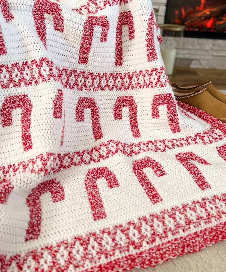 K&ouml;nnte beinhalten: Eine wei&szlig;e geh&auml;kelte Decke mit einem sich wiederholenden Muster aus roten Zuckerstangen. Die Decke hat einen dekorativen roten Rand und ein gemustertes Design zwischen den Zuckerstangen. Perfekt f&uuml;r die Weihnachtszeit.