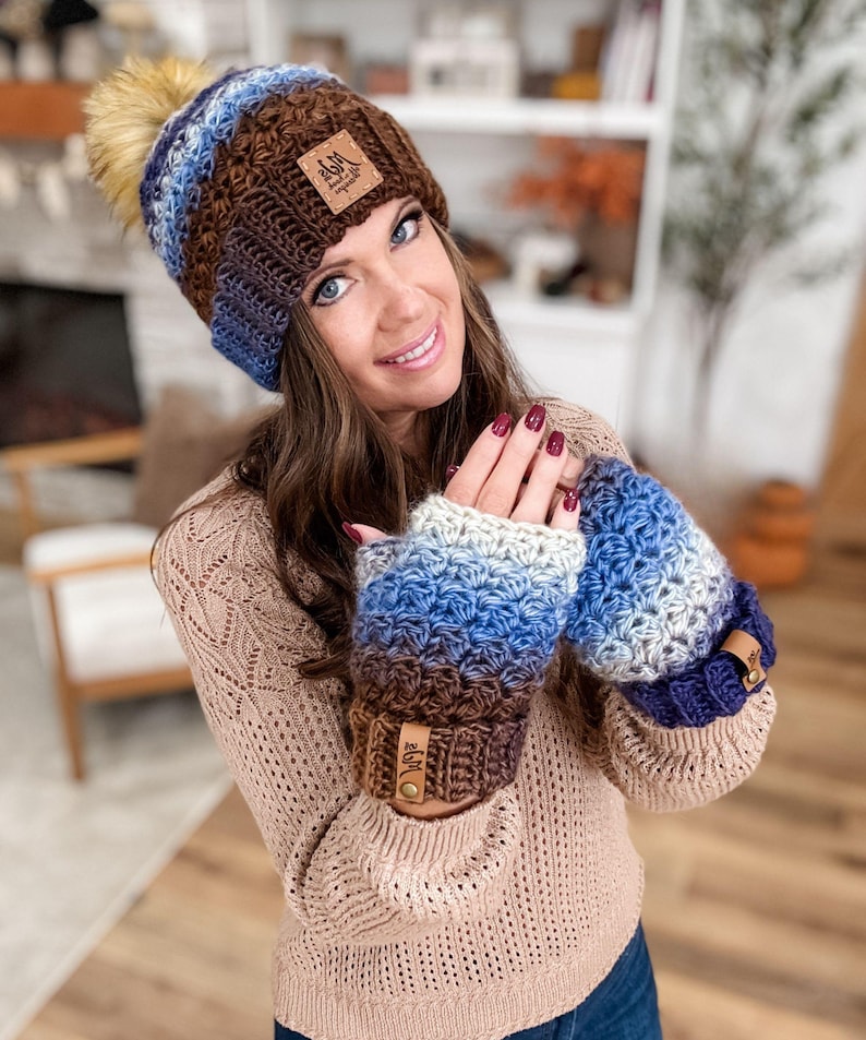 May include: A handmade crochet hat and fingerless gloves set. The hat features a brown, blue, and white color gradient with a faux fur pom-pom. The matching gloves have a similar color scheme and leather tags.