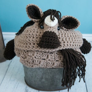 Pu&ograve; includere: Una coperta a forma di cavallo in crochet di colore marrone e bianco con una lunga coda marrone. La coperta &egrave; appoggiata in un secchio di metallo.