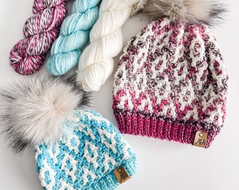 May include: Two handmade knit beanies with faux fur pom-poms, one in blue and white, the other in pink, white, and gray. Several skeins of yarn in coordinating colors are arranged nearby. Each beanie has a small leather tag.
