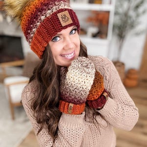 May include: A handmade crochet hat and mittens set in a warm color palette of orange, burgundy, and cream. The hat features a faux fur pom-pom and a leather tag. The mittens have matching wrist cuffs with leather tags.