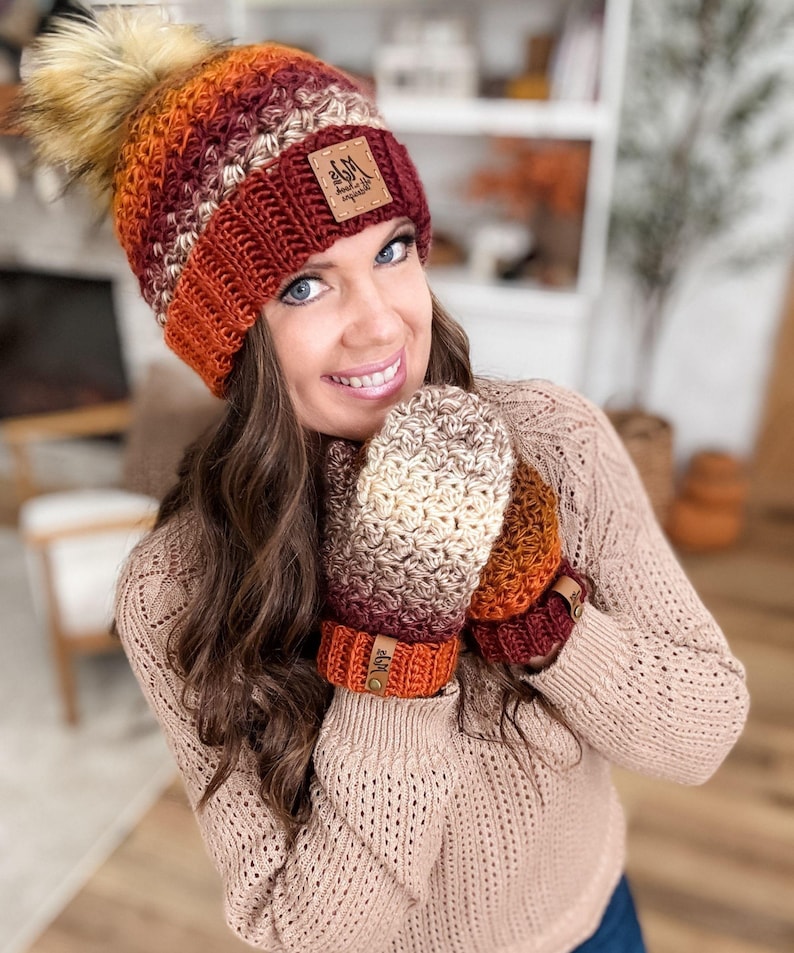 May include: A handmade crochet hat and mittens set in a warm color palette of orange, burgundy, and cream. The hat features a faux fur pom-pom and a leather tag. The mittens have matching wrist cuffs with leather tags.