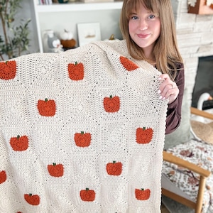 May include: A cream-coloured crochet blanket constructed from square motifs, each featuring a textured orange pumpkin with a small green stem. The blanket is composed of alternating pumpkin squares and plain cream squares, creating a repeating pattern.
