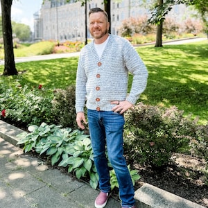 May include: A light blue and white knit cardigan with wooden buttons. The cardigan is worn over a white t-shirt and paired with blue jeans and burgundy sneakers. The background features a park setting with greenery and a building.