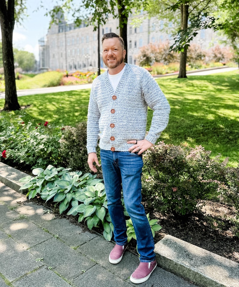 May include: A light blue and white knit cardigan with wooden buttons. The cardigan is worn over a white t-shirt and paired with blue jeans and burgundy sneakers. The background features a park setting with greenery and a building.