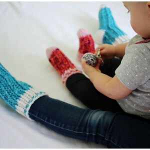 May include: A child wearing blue jeans and a grey shirt with white polka dots sits on a white surface. The child is wearing three pairs of colorful knitted socks. The child is holding a silver toy.