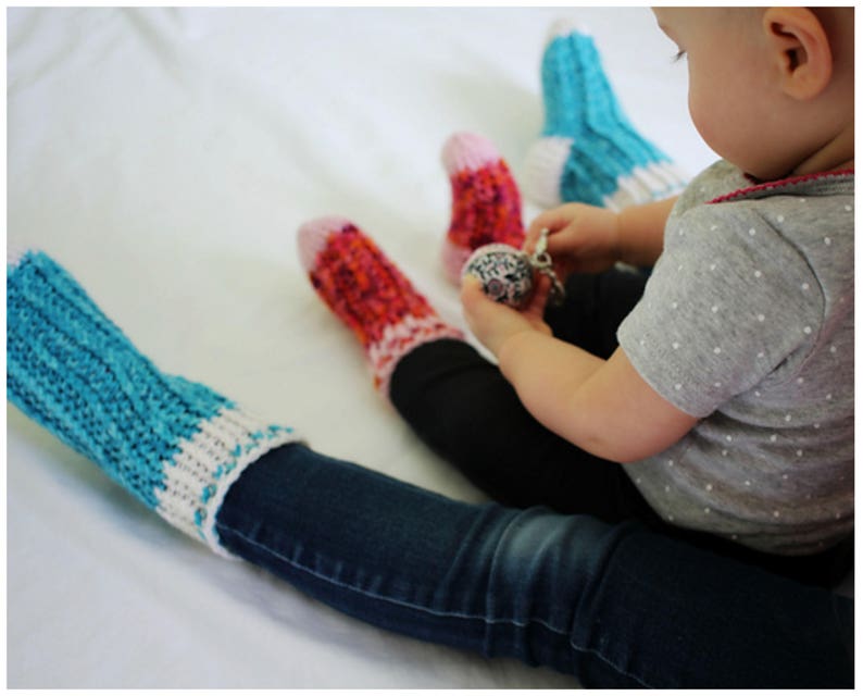 May include: A child wearing blue jeans and a grey shirt with white polka dots sits on a white surface. The child is wearing three pairs of colorful knitted socks. The child is holding a silver toy.