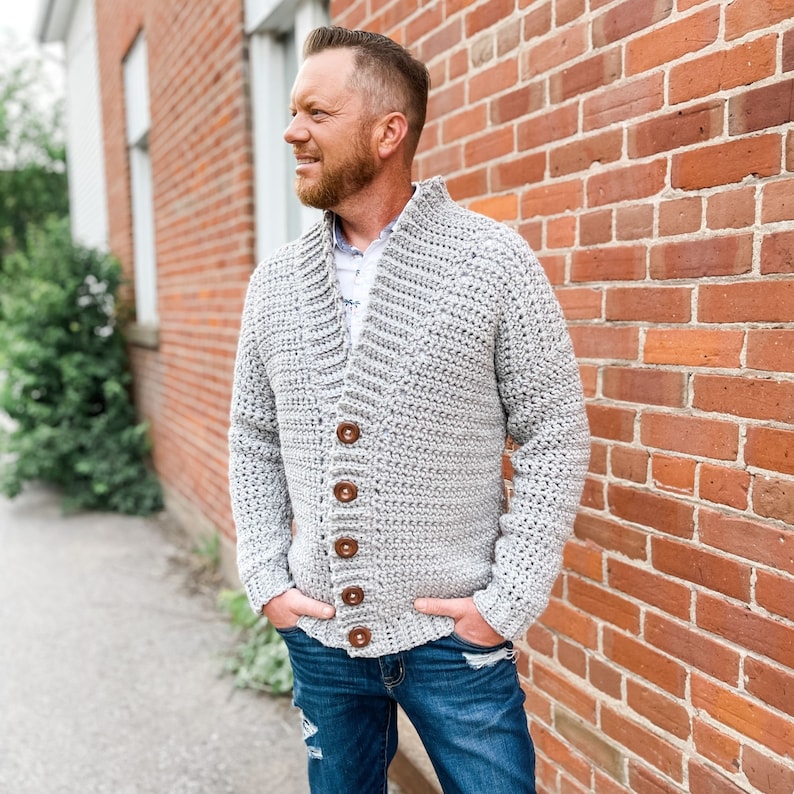 May include: A grey crocheted cardigan with a shawl collar and wooden buttons. The cardigan is being worn by a person standing in front of a brick wall.