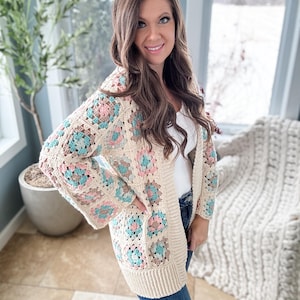 May include: A crocheted cardigan with a granny square pattern in shades of beige, pink, and blue. The cardigan has a long, open front and long sleeves.