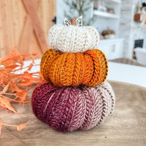 K&ouml;nnte beinhalten: Ein Stapel aus drei gestrickten K&uuml;rbissen in Herbstfarben. Der obere K&uuml;rbis ist cremefarben, der mittlere ist orange und der untere ist eine Mischung aus Burgunderrot und Beige. Ein Holzblatt und -stiel zieren den cremefarbenen K&uuml;rbis. Die K&uuml;rbisse stehen auf einem Holztablett.