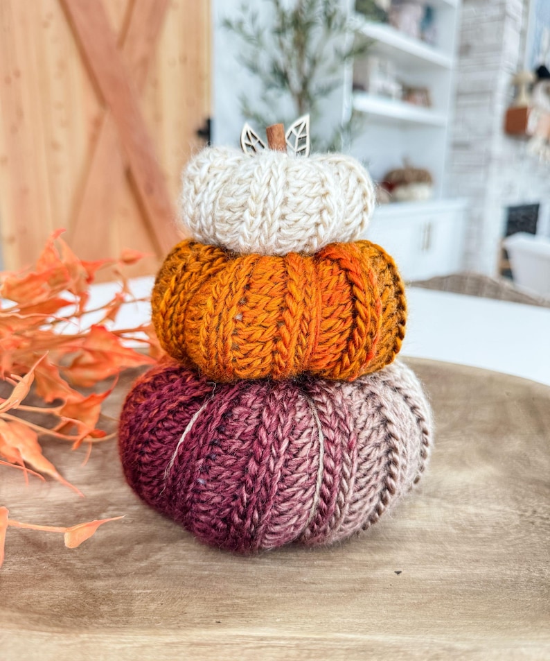 K&ouml;nnte beinhalten: Ein Stapel aus drei gestrickten K&uuml;rbissen in Herbstfarben. Der obere K&uuml;rbis ist cremefarben, der mittlere ist orange und der untere ist eine Mischung aus Burgunderrot und Beige. Ein Holzblatt und -stiel zieren den cremefarbenen K&uuml;rbis. Die K&uuml;rbisse stehen auf einem Holztablett.