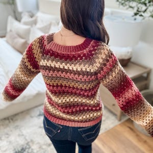 Pu&ograve; includere: Un maglione all'uncinetto multicolore nei toni del bordeaux, marrone e beige. Il maglione ha uno scollo quadrato e maniche lunghe. La persona indossa jeans blu scuro. Il maglione &egrave; fatto a mano e ha un aspetto strutturato.
