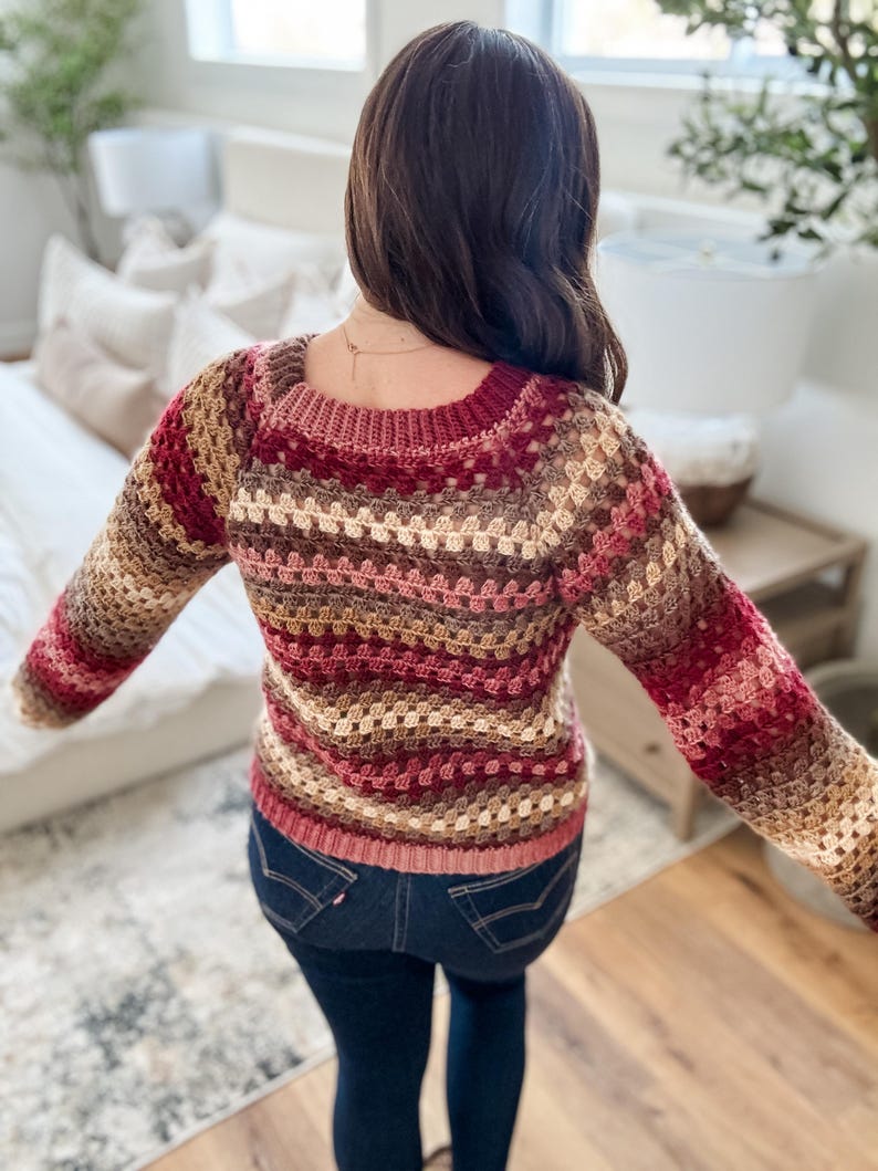 Pu&ograve; includere: Un maglione all'uncinetto multicolore nei toni del bordeaux, marrone e beige. Il maglione ha uno scollo quadrato e maniche lunghe. La persona indossa jeans blu scuro. Il maglione &egrave; fatto a mano e ha un aspetto strutturato.