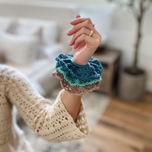 May include: A stack of three crocheted scrunchies in shades of teal, green, and brown. The scrunchies are handmade and have a ruffled edge. The arm is wearing a cream-colored crocheted sweater.