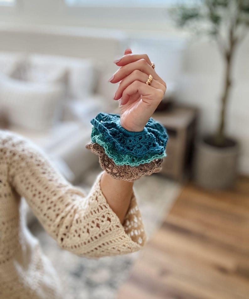 May include: A stack of three crocheted scrunchies in shades of teal, green, and brown. The scrunchies are handmade and have a ruffled edge. The arm is wearing a cream-colored crocheted sweater.
