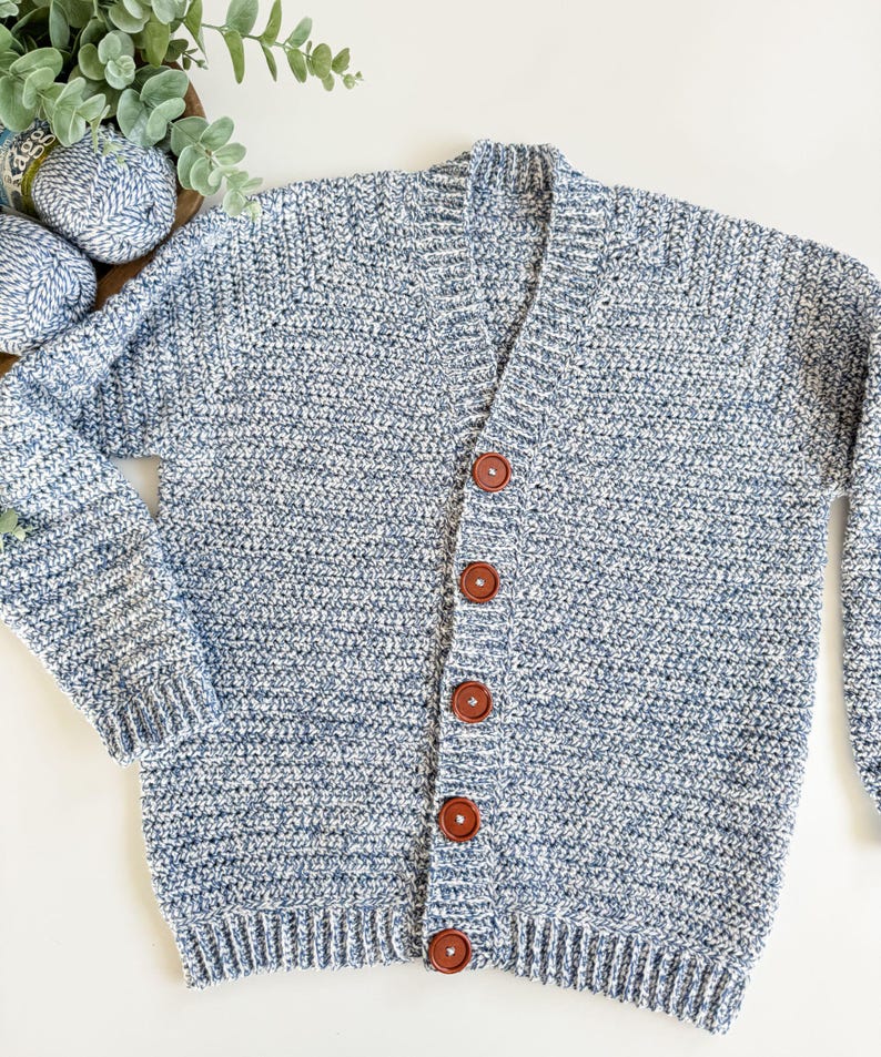 May include: A blue and white speckled knit cardigan with a V-neck and long sleeves. The sweater has five brown buttons down the front. The knitwear is displayed on a white surface.