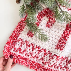 K&ouml;nnte beinhalten: Ein handgefertigter, geh&auml;kelter Weihnachtsbaumrock in Rot und Wei&szlig;. Der Rock hat ein sich wiederholendes Muster aus roten Streifen auf wei&szlig;em Hintergrund mit einem geschwungenen roten Rand. Gr&uuml;ne Kiefernzweige und ein Tannenzapfen befinden sich in der oberen linken Ecke.