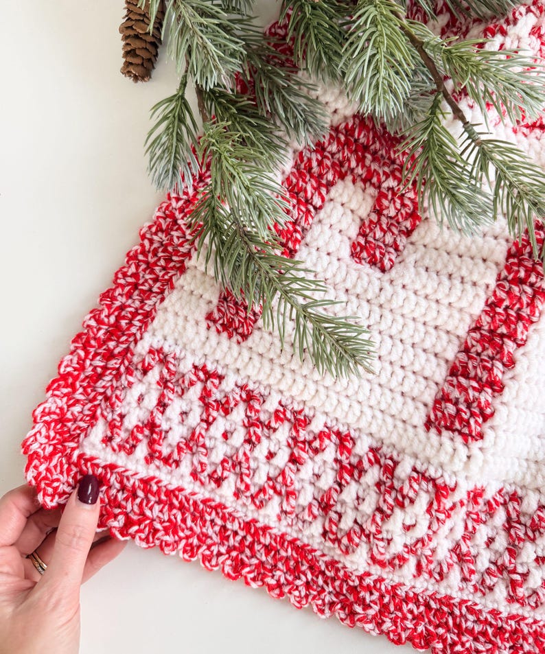 K&ouml;nnte beinhalten: Ein handgefertigter, geh&auml;kelter Weihnachtsbaumrock in Rot und Wei&szlig;. Der Rock hat ein sich wiederholendes Muster aus roten Streifen auf wei&szlig;em Hintergrund mit einem geschwungenen roten Rand. Gr&uuml;ne Kiefernzweige und ein Tannenzapfen befinden sich in der oberen linken Ecke.
