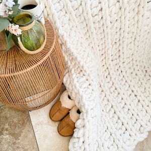Peut inclure: Une couverture en tricot blanc &eacute;paisse drap&eacute; sur une table d'appoint en osier tress&eacute; avec un vase en verre et une paire de pantoufles en fausse fourrure marron.