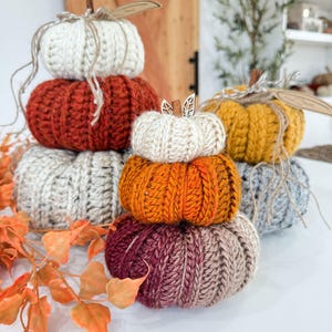 Puede incluir: Una colección de calabazas hechas a mano, apiladas, en varios colores, incluyendo blanco, naranja, óxido y gris. Cada calabaza está hecha de hilo de punto grueso, con un tallo de ramita y cuerda. Artículos decorativos con temática otoñal.
