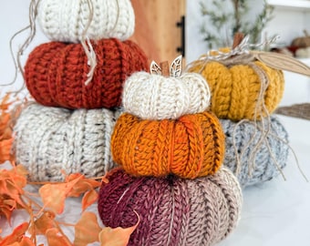 Puede incluir: Una colección de calabazas hechas a mano, apiladas, en varios colores, incluyendo blanco, naranja, óxido y gris. Cada calabaza está hecha de hilo de punto grueso, con un tallo de ramita y cuerda. Artículos decorativos con temática otoñal.