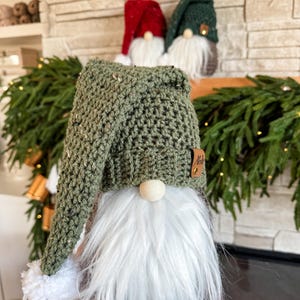 May include: A green crocheted gnome with a white beard and a white pom-pom on the hat. The gnome is sitting in front of a fireplace.