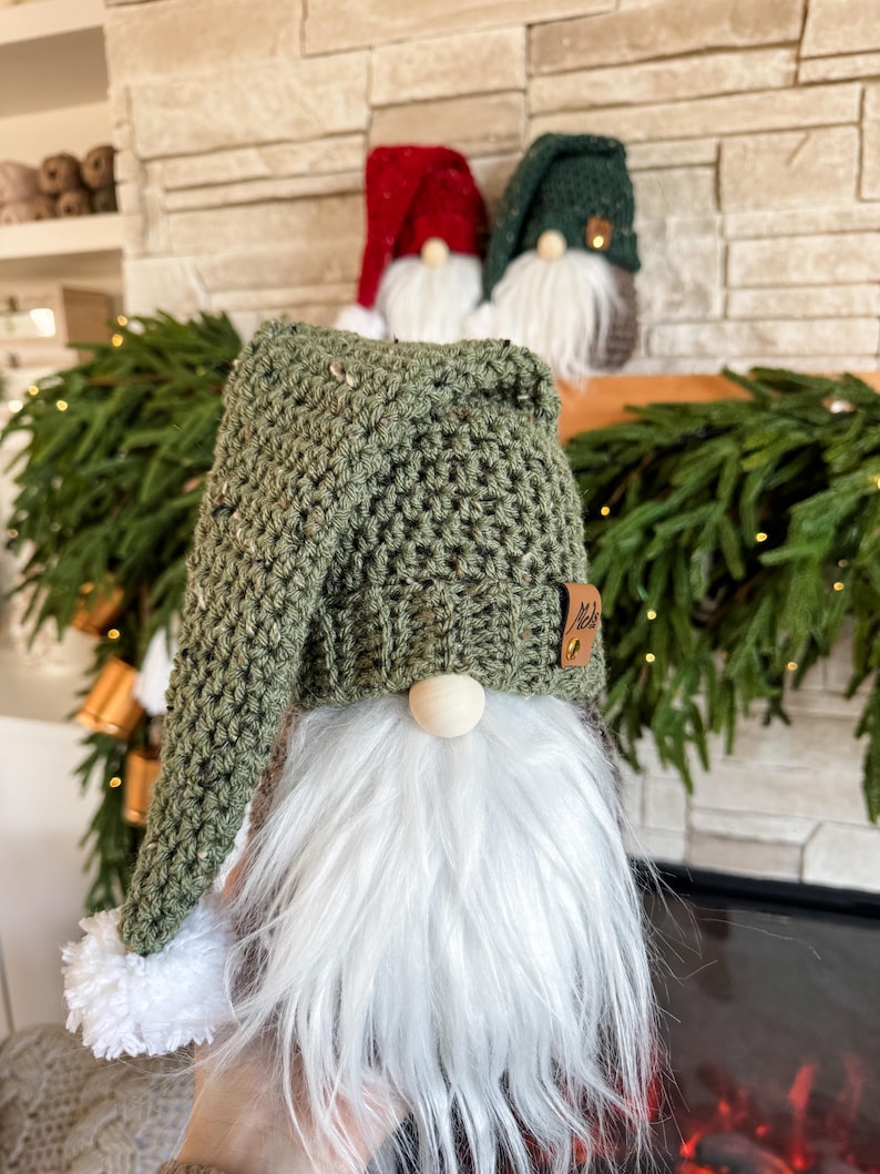 May include: A green crocheted gnome with a white beard and a white pom-pom on the hat. The gnome is sitting in front of a fireplace.