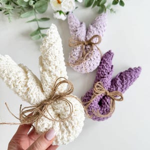 May include: Three crocheted bunny shapes, one white, one light purple, and one dark purple. Each bunny has a brown twine bow tied around its neck.
