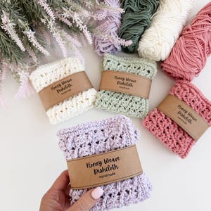 Puede incluir: Pa&ntilde;os de cocina Honey Weave hechos a mano en crema, verde salvia, rosa y lavanda, cada uno envuelto con una banda de papel marr&oacute;n. Los pa&ntilde;os de cocina se exhiben con madejas de hilo en colores coordinados. El texto en la banda dice "Honey Weave Dishcloth Handmade."