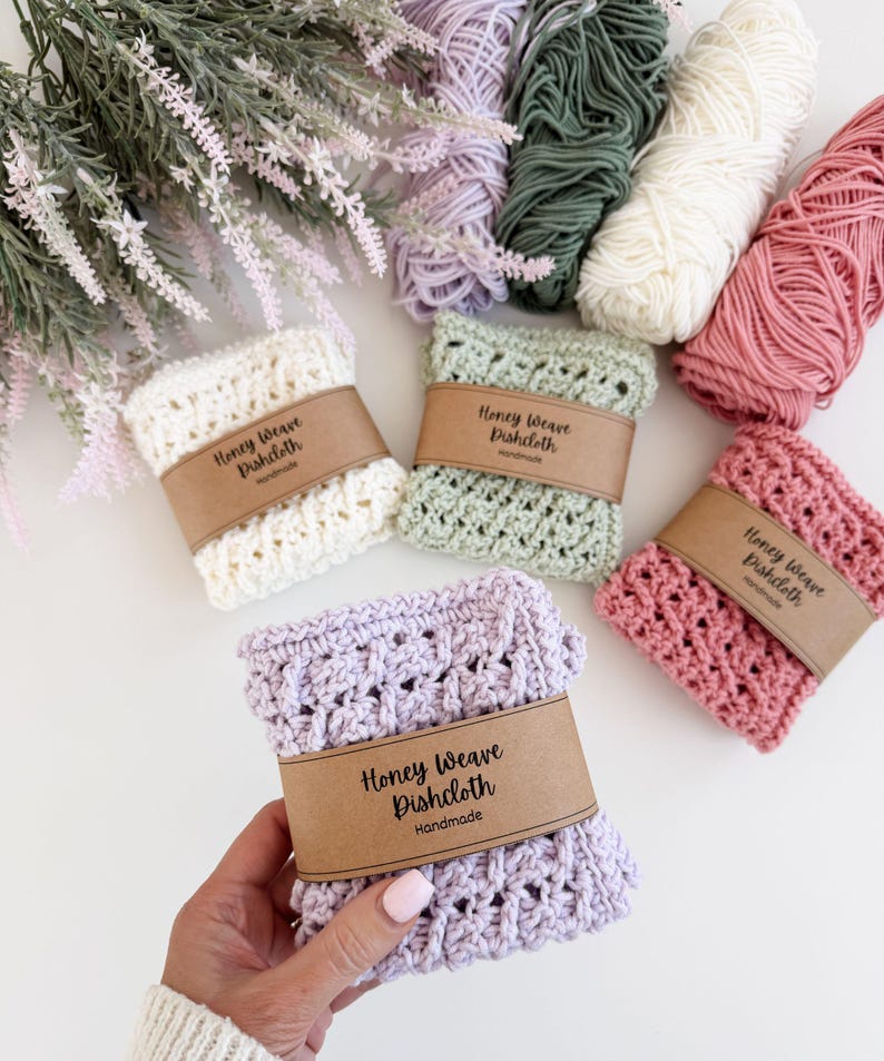 Puede incluir: Pa&ntilde;os de cocina Honey Weave hechos a mano en crema, verde salvia, rosa y lavanda, cada uno envuelto con una banda de papel marr&oacute;n. Los pa&ntilde;os de cocina se exhiben con madejas de hilo en colores coordinados. El texto en la banda dice "Honey Weave Dishcloth Handmade."