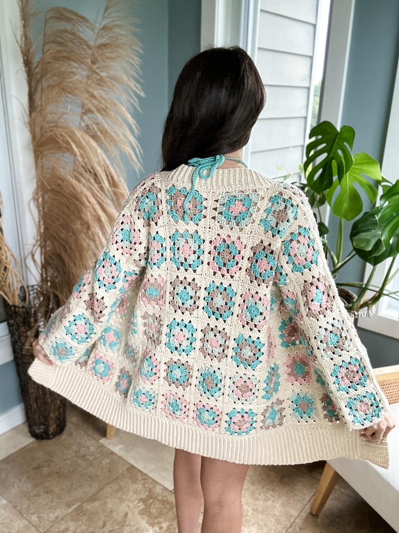 May include: A crocheted cardigan with a square pattern in shades of pink, blue, and brown. The cardigan has a loose fit and long sleeves.
