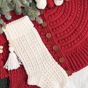 May include: A cream-colored, textured, crocheted Christmas stocking with a white tassel and wooden beads. The stocking is placed on a red crocheted tree skirt with wooden buttons. Green pine branches and yarn balls are in the background.