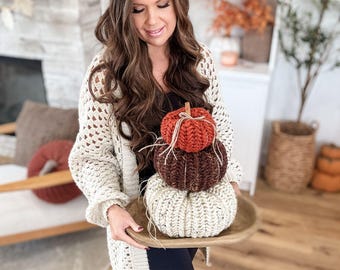 May include: A wooden tray holds three handmade, knitted pumpkins in shades of orange, brown, and cream. The pumpkins are stacked, with twine and a small twig accenting the top pumpkin. The pumpkins are a fall-themed decorative item.