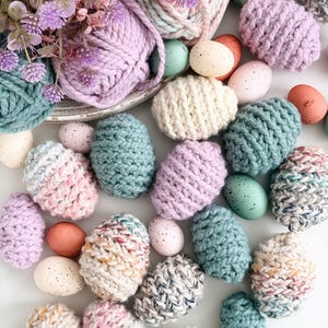 May include: An assortment of colorful, knitted Easter eggs in shades of blue, pink, purple, and cream. Speckled and solid-colored eggs are scattered around yarn and faux flowers, creating a festive spring display.