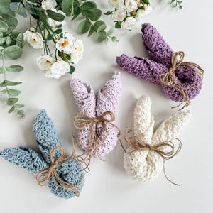 May include: Three crocheted bunny shapes in lavender, white, and blue, tied with twine. The bunnies are on a white surface.